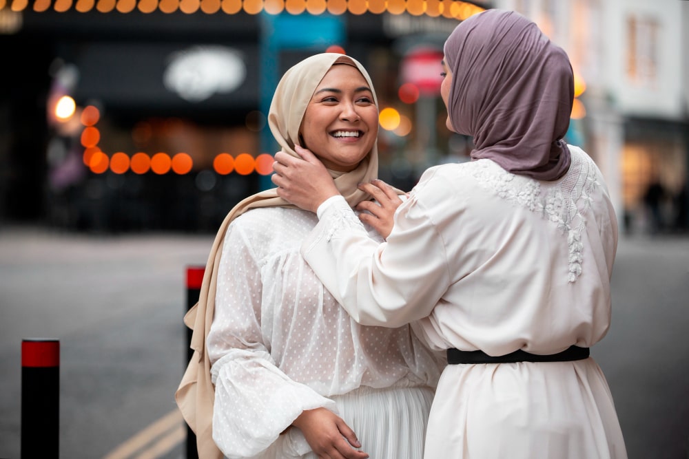 Ilustrasi dua wanita muslimah tersenyum, salah satunya membantu membenahi hijab temannya sebagai simbol menutupi aib sahabat dalam adab berteman menurut Islam